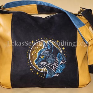 Könnte beinhalten: Große, schwarze Umhängetasche mit Katzenmotiv. Die Tasche hat eine schwarze Wildleder-Mittelbahn mit einem gestickten Katzenprofil in einem Kreis, akzentuiert mit Gold und Blau. Die Seiten sind aus senfgelbem Leder mit blauen und schwarzen Riemen.