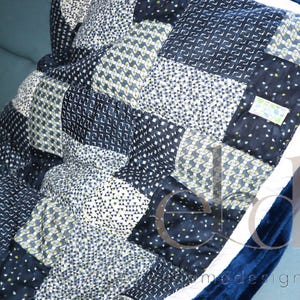 Könnte beinhalten: Eine Patchwork-Decke mit verschiedenen dunkelblauen, marineblauen und weißen geometrischen Mustern in quadratischer Anordnung. Die Decke ist mit einem dunkelblauen, samtartigen Stoff eingefasst und weist ein kleines rechteckiges Etikett auf.