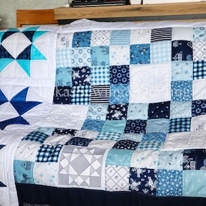 Könnte beinhalten: Eine handgefertigte Steppdecke in Blau- und Weißtönen mit Stern- und geometrischen Mustern. Die Steppdecke besteht aus verschiedenen Stoffquadraten, die ein Patchwork-Design bilden. Die Steppdecke wird auf einer dunkelblauen Oberfläche präsentiert.