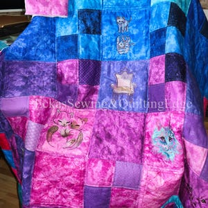 Könnte beinhalten: Eine Patchwork-Decke aus Stoffquadraten in verschiedenen Blau-, Lila- und Rosatönen. Die Decke enthält gestickte Katzenmotive, darunter Katzen, die auf einem Stuhl ruhen und zusammen schlafen. Der Text EckasSewing&QuiltingEdge ist sichtbar.