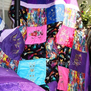 Può includere: Una trapunta patchwork con vari quadrati di tessuto a tema gatto in tonalità viola, rosa, blu e nero. La trapunta presenta disegni di gatti ricamati, accenti floreali e il testo TexasSewing&QuiltingEdge stampato al centro del design.