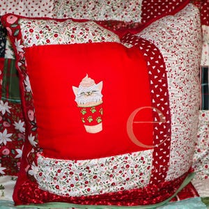 Könnte beinhalten: Ein quadratisches Zierkissen mit einem zentralen, einfarbig roten Feld, das eine Stickerei einer Katze in einer Tasse mit Sahnehäubchen zeigt. Das Kissen ist von Patchwork-Stoffmustern in roten, weißen und grünen Blumenmotiven umrandet.
