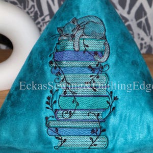 Könnte beinhalten: Ein türkisfarbenes, dreieckiges Kissen mit einem gestickten Design. Eine hellblaue Katze schläft auf einem Stapel Bücher in Blau- und Türkistönen. Das Design ist mit schwarzen Ranken und dem Text "EckasSewing&QuiltingEdge" akzentuiert.