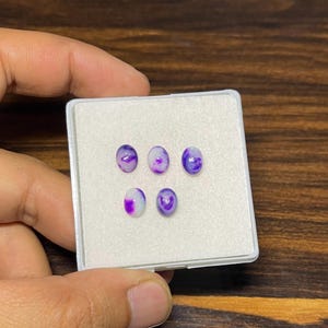 May include: A small, white plastic container holds six oval-shaped gemstones. The gemstones are a mix of purple and white, with a marbled appearance. The container is held by a hand, and the background is a dark wood surface.