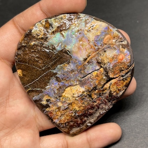 Può includere: Un esemplare di opale lucido a forma di goccia. La pietra presenta una matrice marrone scuro con colori interni vibranti tra cui blu, verdi e arancioni. La superficie presenta crepe e variazioni naturali, mostrando il carattere unico dell'opale.