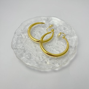 May include: A pair of gold hoop earrings with clip-on backs. The earrings are on a white, textured surface.