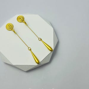 Op de afbeelding: Een paar gouden oorhangers met een cirkelvormig ontwerp aan de bovenkant en een traanvorm aan de onderkant. De oorhangers liggen op een wit oppervlak.