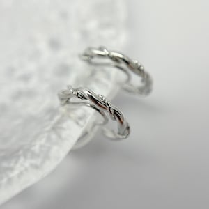 Könnte beinhalten: Ein Paar silberne, gedrehte Creolen. Die Ohrringe bestehen aus einem dünnen, gedrehten Draht und haben ein schlichtes, elegantes Design.