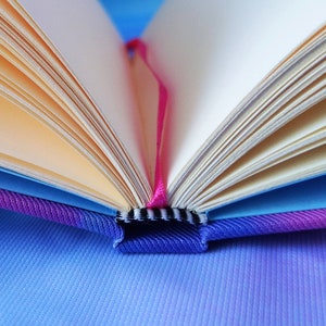 Op de afbeelding: Een close-up van een boek met een roze lint als bladwijzer. Het boek is open en de pagina's zijn uitwaaierend. Het boek is gebonden in een blauwe en paarse stoffen omslag.