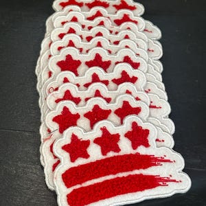 May include: A stack of white and red embroidered patches. Each patch features a red star design above two red stripes, all outlined in white. The patches are likely for decorative purposes, such as for clothing or accessories.