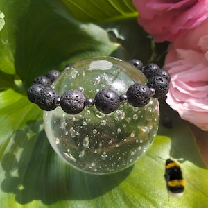 Peut inclure: Un bracelet de perles en pierre de lave noire avec des perles d'accent argentées. Le bracelet est présenté sur une sphère de verre transparente, entourée de feuilles vertes et de fleurs roses. Les perles de lave ont une texture poreuse.