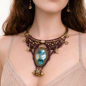 Puede incluir: Un collar artesanal con un gran colgante ovalado de piedra preciosa azul y verde. El colgante está engastado en un marco de color bronce y está rodeado de intrincado trabajo de macramé en tonos marrón y dorado. Pendientes a juego.
