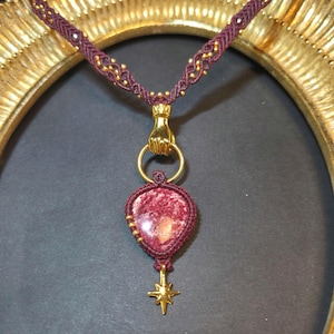 May include: A necklace featuring a dark maroon macrame cord with small gold beads, a gold-toned hand clasp holding a circular ring, a teardrop-shaped red stone pendant encased in maroon macrame, and a small gold-toned star charm at the bottom.