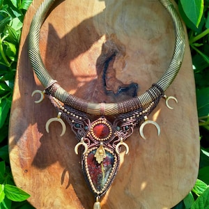 Puede incluir: Un collar artesanal con un collar de tono latón con un diseño texturizado. El colgante presenta una piedra roja y marrón, acentuada con dijes de media luna dorados y una borla dorada. El collar se exhibe sobre una superficie de madera.