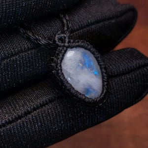 Puede incluir: Un collar con colgante hecho a mano con una piedra lunar pulida de forma ovalada. La piedra lunar está engastada en un diseño de macramé negro y cuelga de un cordón negro a juego. La piedra muestra un sutil juego de colores azules y blancos.