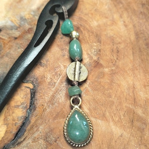 Puede incluir: Una horquilla de madera negra con un adorno de cuentas decorativo. El adorno presenta cuentas de piedra verde, un disco de color dorado y un colgante de piedra verde en forma de lágrima con un borde dorado en forma de cuerda. El accesorio para el cabello se muestra sobre una superficie de madera.