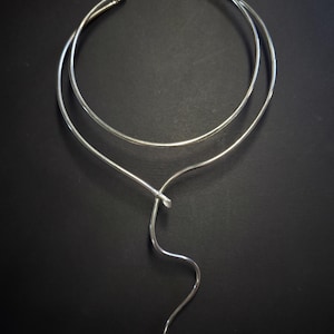 Könnte beinhalten: Eine silberfarbene Metall-Halskette in Choker-Form mit einem kreisförmigen Band, das sich vorne überlappt und in einen einzelnen, schlanken, wellenförmigen Ausläufer übergeht, der nach unten schwingt. Das Stück hat eine glatte, polierte Oberfläche.