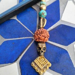 Puede incluir: Un colgante artesanal con un dije de calavera y cuentas decorativas. El colgante tiene una calavera de color dorado y un dije en forma de diamante. Las cuentas incluyen elementos turquesa, dorados y marrones, ensartados en un cordón marrón.