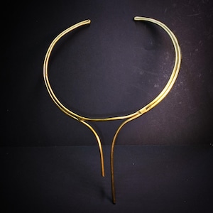 May include: A gold-colored necklace with a minimalist design. The necklace features a circular upper portion and two vertical bars that hang down. The jewelry is set against a dark background.