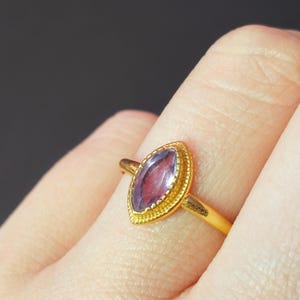 Puede incluir: Un anillo de oro con una piedra preciosa púrpura en forma de marquesa. La piedra preciosa está engastada en un bisel detallado y texturizado. La banda del anillo es de oro pulido. El anillo se muestra en un dedo.