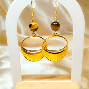 Puede incluir: Un par de pendientes dorados con un diseño circular. Cada pendiente presenta un colgante dorado en forma de media luna y una cuenta redonda marrón y amarilla. Los pendientes se exhiben en un soporte blanco con una base de madera.