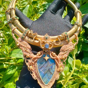 May include: A handcrafted statement necklace featuring a large, blue labradorite pendant. The necklace has a gold-tone metal collar and intricate macrame detailing in shades of tan and gold. Accented with green beads and a sunburst design.
