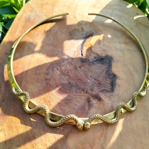May include: A gold-coloured metal choker necklace. The necklace features a snake design with detailed scales. The snake wraps around the neckpiece, with the snake heads meeting at the front. The necklace is displayed on a wooden surface.