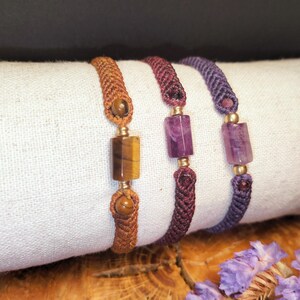 Puede incluir: Tres pulseras de macramé con cuentas de piedras preciosas. Una pulsera presenta una piedra de ojo de tigre, las otras dos tienen piedras de amatista. Las pulseras son en tonos marrón, burdeos y morado, con detalles dorados.
