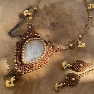 Puede incluir: Un conjunto de collar y pendientes de macramé hechos a mano. El collar presenta una gran piedra ovalada de color claro engastada en un diseño de macramé marrón, acentuado con cuentas y dijes dorados. Los pendientes tienen un diseño similar.