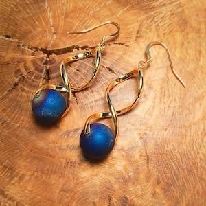 Puede incluir: Un par de pendientes dorados con un diseño en espiral. Cada pendiente presenta una cuenta esférica azul oscuro. Los pendientes se exhiben sobre una superficie de madera, mostrando su elegante diseño y contraste de color.