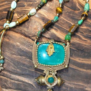 Puede incluir: Collar hecho a mano con un colgante de piedra turquesa y un dije de mano de Hamsa dorado. El collar incorpora cuentas verdes, marrones y doradas en un cordón. El colgante mide aproximadamente 5 cm de ancho.