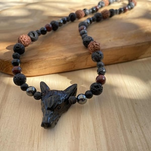 Puede incluir: Un collar de cuentas con un colgante de lobo negro tallado. El collar incorpora varias cuentas en tonos negros, marrones y grises, incluyendo roca volcánica y hematita. El collar se exhibe sobre una superficie de madera.