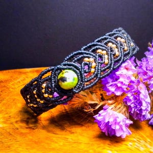 Puede incluir: Una pulsera de macramé negra con una piedra central verde y amarilla y cuentas doradas. La pulsera se muestra sobre una superficie de madera, con flores moradas en el fondo. El intrincado anudado de la pulsera es visible.