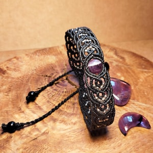Puede incluir: Una pulsera de macramé negra con una piedra preciosa púrpura central y cuentas plateadas. La pulsera se exhibe sobre una superficie de madera, con dos piedras preciosas púrpuras adicionales cerca. La pulsera tiene cordones negros ajustables.