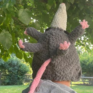 May include: A grey crochet opossum hat with a white cone-shaped head, pink paws, and a pink tail. The hat is designed to be worn on the head, with the opossum's features extending outwards.