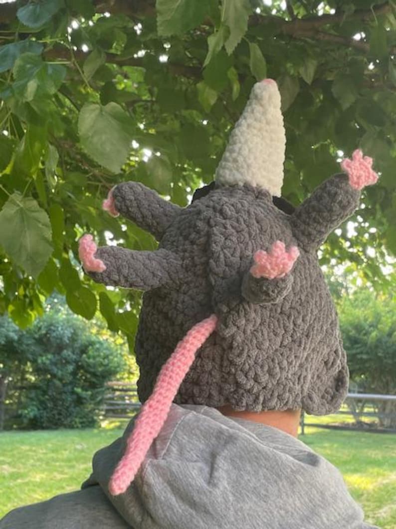May include: A grey crochet opossum hat with a white cone-shaped head, pink paws, and a pink tail. The hat is designed to be worn on the head, with the opossum's features extending outwards.