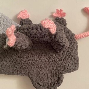 May include: A handmade crochet opossum hat in grey, light blue, and pink. The hat features a grey body, light blue ears, pink paws, and a pink tail. The hat is designed to be worn on the head.