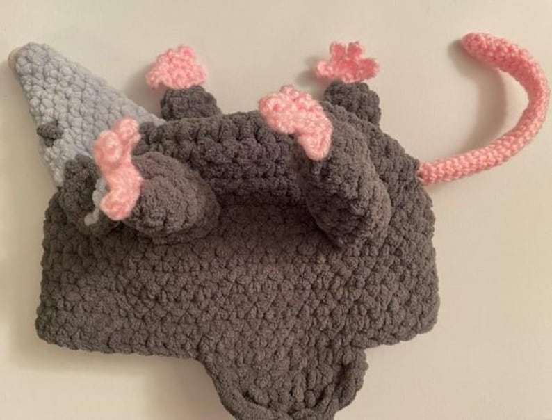 May include: A handmade crochet opossum hat in grey, light blue, and pink. The hat features a grey body, light blue ears, pink paws, and a pink tail. The hat is designed to be worn on the head.