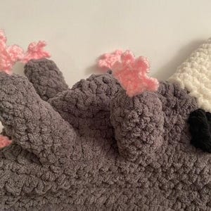 May include: A handmade crochet bird hat. The hat is mainly grey with a white beak, black details, and pink accents. The hat is made of a soft, textured yarn, perfect for a child.