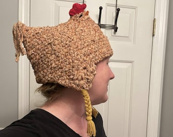 May include: A brown crocheted chicken hat with a red comb and yellow yarn braids. The hat is being worn by a person.