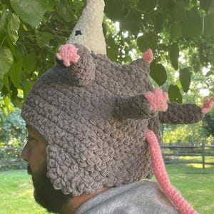 May include: A grey crocheted hat in the shape of a possum, with a white nose, pink ears and paws, and a pink tail. The hat is worn by a person. This novelty hat is a fun and quirky accessory.