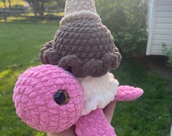 May include: A plush crochet turtle toy featuring a pink head and flippers, a white underbelly, and a brown textured shell shaped like a scoop of ice cream topped with a beige cone. The turtle has a single visible dark eye with gold glitter accents.