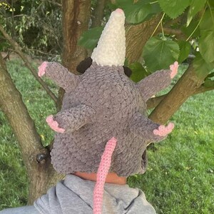 May include: A grey crocheted opossum hat with a white snout, pink ears, and a pink tail. The hat is made of a soft, textured yarn and is designed to fit over the head. The hat has a whimsical and playful design.
