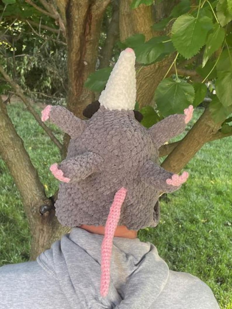 May include: A grey crocheted opossum hat with a white snout, pink ears, and a pink tail. The hat is made of a soft, textured yarn and is designed to fit over the head. The hat has a whimsical and playful design.