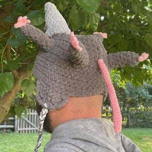 May include: A grey crocheted hat with a white horn, pink ears, and a long pink tail. The hat has a braided cord with grey and white strands. The hat is designed to resemble a rat.
