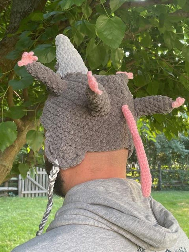 May include: A grey crocheted hat with a white horn, pink ears, and a long pink tail. The hat has a braided cord with grey and white strands. The hat is designed to resemble a rat.