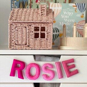 Puede incluir: Una cesta de almacenamiento de mimbre rosa en forma de casa se encuentra en un estante blanco con una pancarta de letras de fieltro que dice "ROSIE" en rosa y rojo.