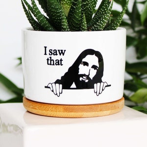 May include: White ceramic planter with a black and white image of Jesus peeking over the edge and the text "I saw that". A green succulent plant is growing in the planter.
