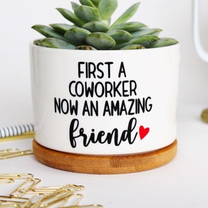 May include: A white ceramic planter with a succulent plant. The planter has the text "FIRST A COWORKER NOW AN AMAZING friend" in black, with a red heart. It sits on a round bamboo coaster. Gold paper clips and a pen are in the background.