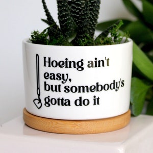 May include: White ceramic planter with a wood base. The planter has a black graphic that says "Hoeing ain't easy, but somebody's gotta do it".